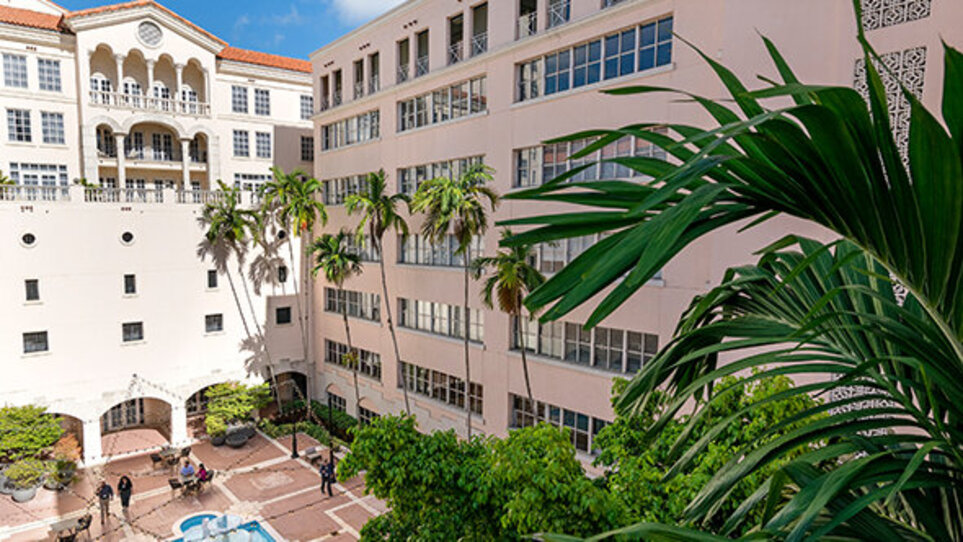 A white seven-storey building with a red roof overlooks a large pink-tiled courtyard with a central pond, green shrubs and seating areas on Kellogg-Miami campus
