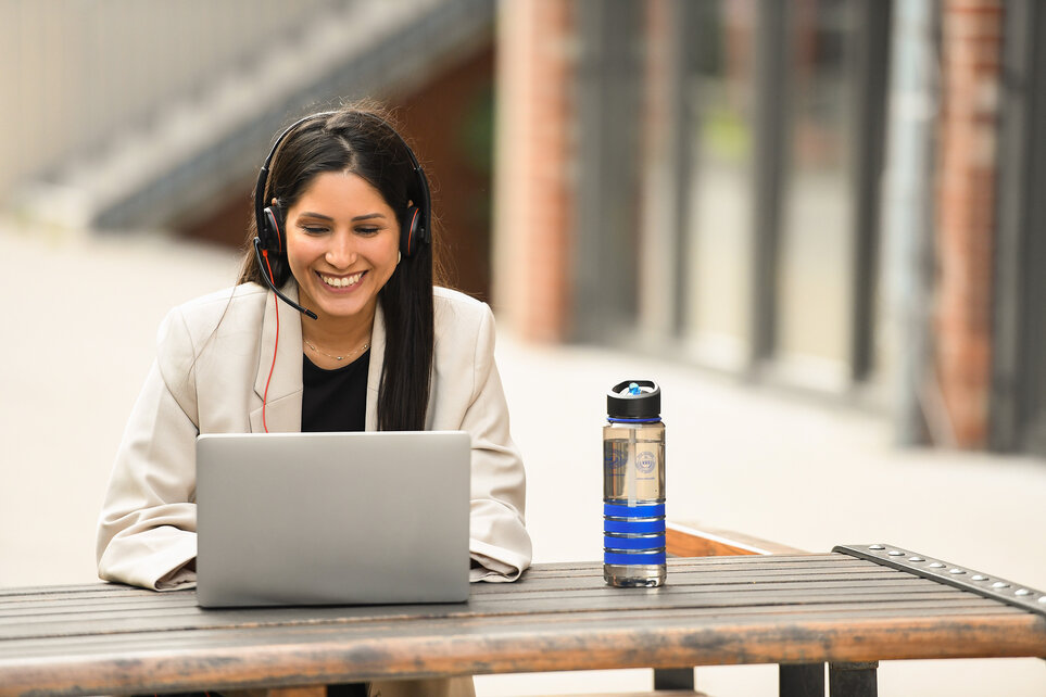A young woman with long black hair, wearing a white blazer and oversized headphones, is sitting outside at a wooden table, working on her laptop.