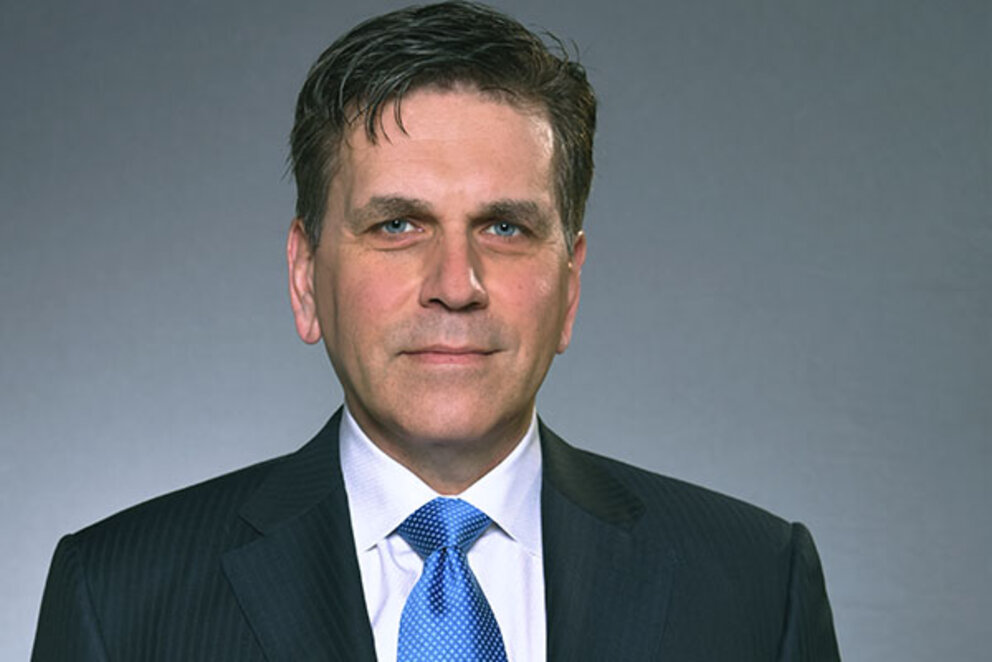 A dark-haired man in a suit looks directly into the camera