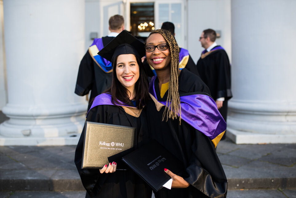 Zwei Absolventinnen in Graduierungsroben mit violetten Schärpen posieren auf Stufen vor zwei großen weißen Säulen und lächeln in die Kamera.