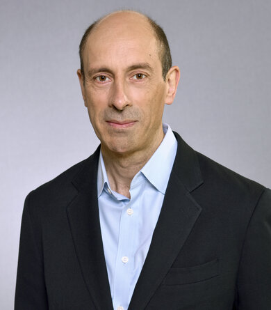 a balding middle-aged man wearing a dark suit and an open-necked blue shirt looks into the camera