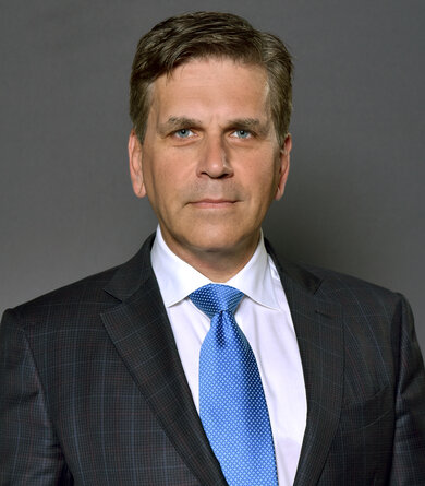 A dark-haired man in a suit looks directly into the camera