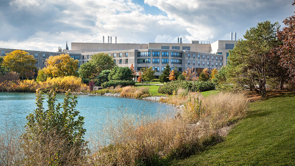 A view of the sprawling grey buildings of Kellogg Evanston campus with a lake and green parkland in the foreground