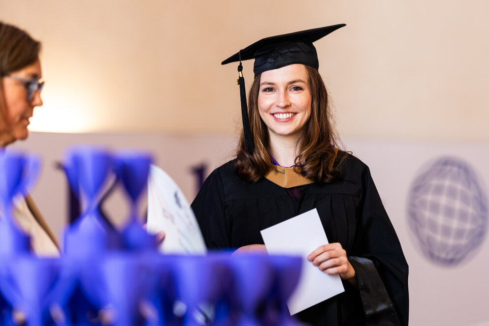 Eine Absolventin in schwarzer Abschlussrobe und Kappe steht hinter einem Tisch mit blauen Miniaturtrophäen und hält stolz eine Urkunde in der Hand.
