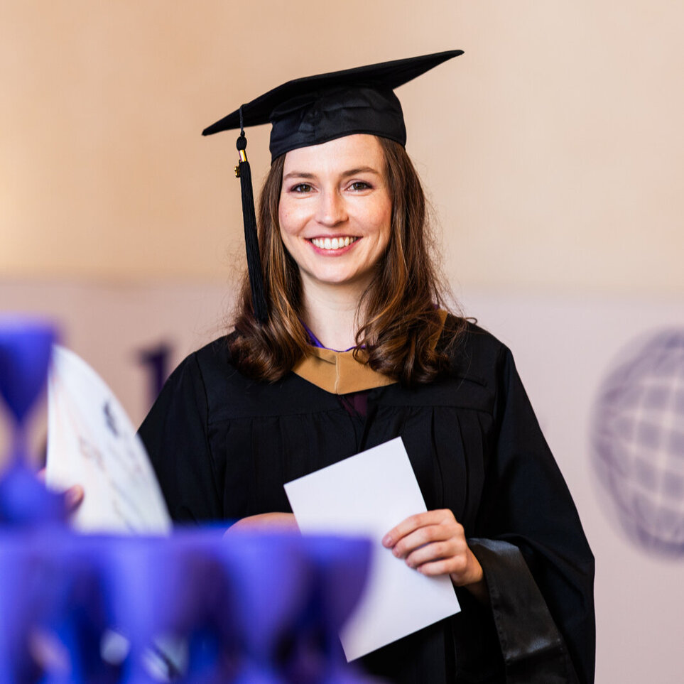 A female graduate wearing a black graduation robe and cap stands behind a table holding miniature blue trophies and proudly holds a certificate.