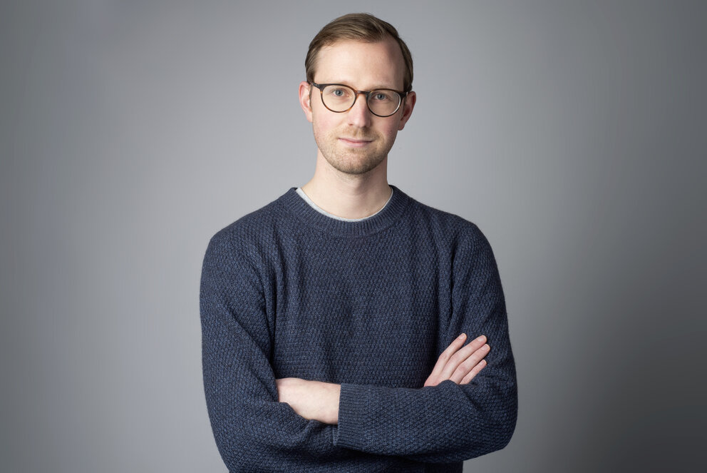 A young man in glasses and a grey sweater poses with his arms crossed
