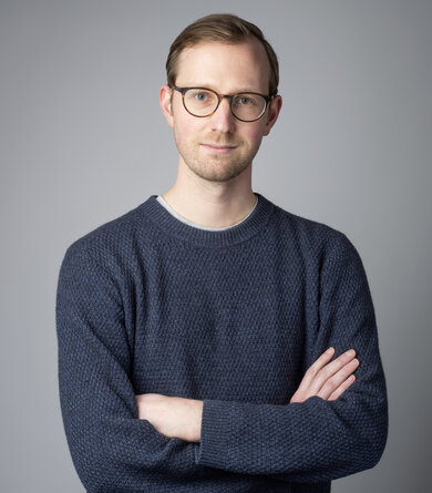 A young man in glasses and a grey sweater poses with his arms crossed