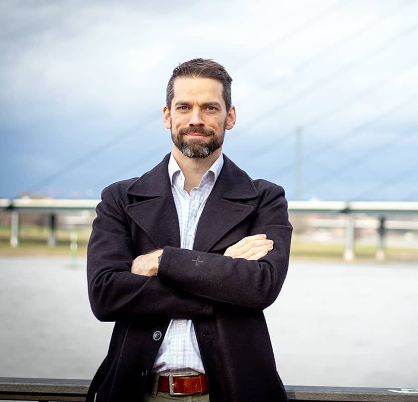 A dark-haired man with a beard, wearing a black jacket, poses with folded arms in front of a suspension bridge.