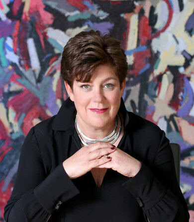 A woman with short dark hair wearing a black blouse sits in front of a modern abstract art painting and stares into the camera