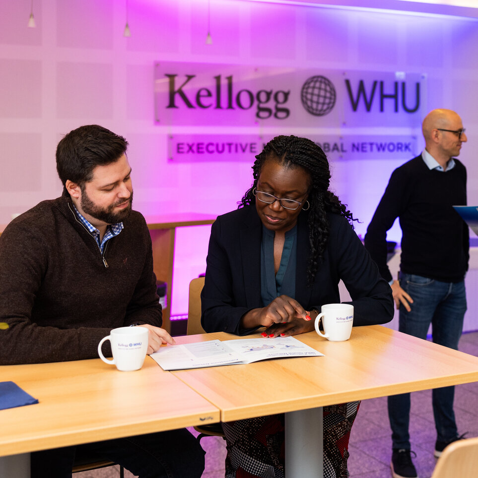 Ein Mann und eine Frau sitzen an einem Tisch, trinken Kaffee und schauen sich eine Broschüre an. Zwei Männer stehen hinter ihnen und unterhalten sich vor einem Schild an der Wand, auf dem "Kellogg-WHU Executive MBA Network" steht.