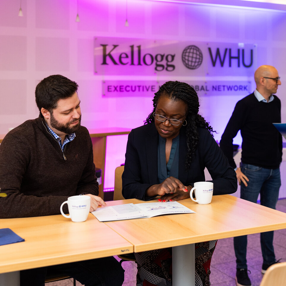 A man and a women sit at a table drinking coffee and looking at a brochure. Two men stand behind them chatting in front of a sign on the wall which says Kellogg-WHU Executive MBA Network.