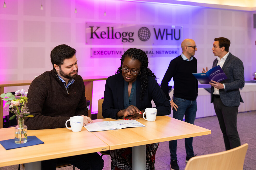 A man and a women sit at a table drinking coffee and looking at a brochure. Two men stand behind them chatting in front of a sign on the wall which says Kellogg-WHU Executive MBA Network.