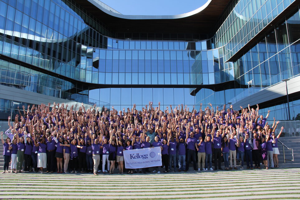 A large group in purple shirts cheers in front of a modern glass building, holding a banner