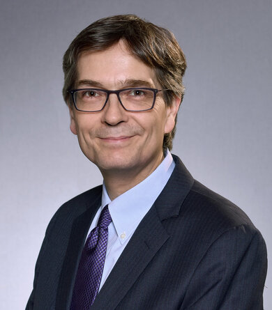 A dark blond man wearing a shirt, tie and dark suuit and glasses smiles into the camera