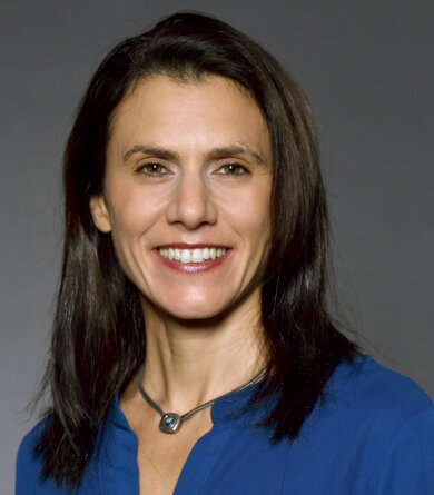 A woman with dark hair and a blue blouse looks into the camera