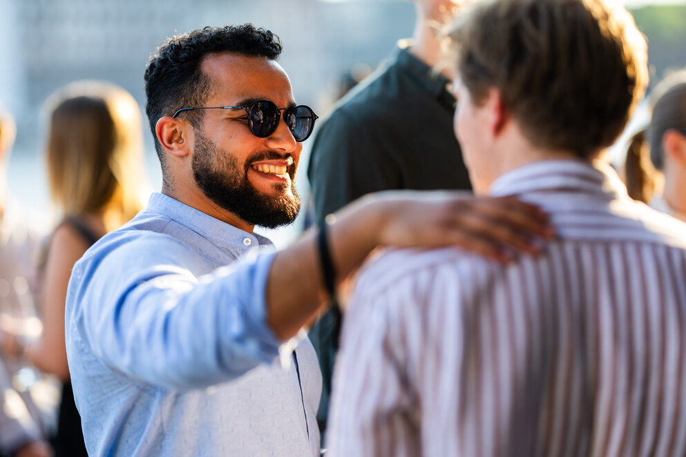 Ein bärtiger dunkelhaariger Mann mit blauem Hemd und Sonnenbrille begrüßt einen anderen Mann, dem er die Hand auf die Schulter legt, bei einem Treffen im Freien.