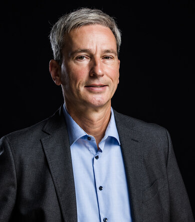 A grey-haired man wearing a blue shirt and dark suit jacket looks into the camera