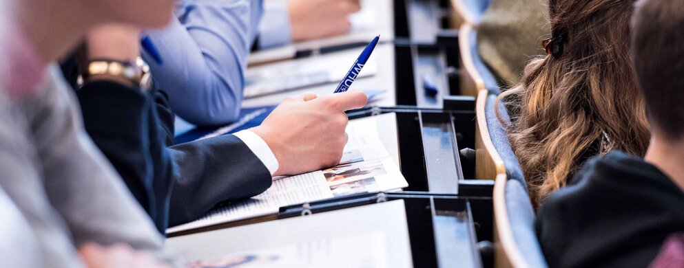 Eine Person sitzt an einem Tisch und hält einen Stift mit der Aufschrift "WHU" in der Hand.