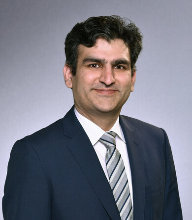 A man with dark wavy hair wearing a navy suit with a white shirt and striped tie looks friendlily into the camera